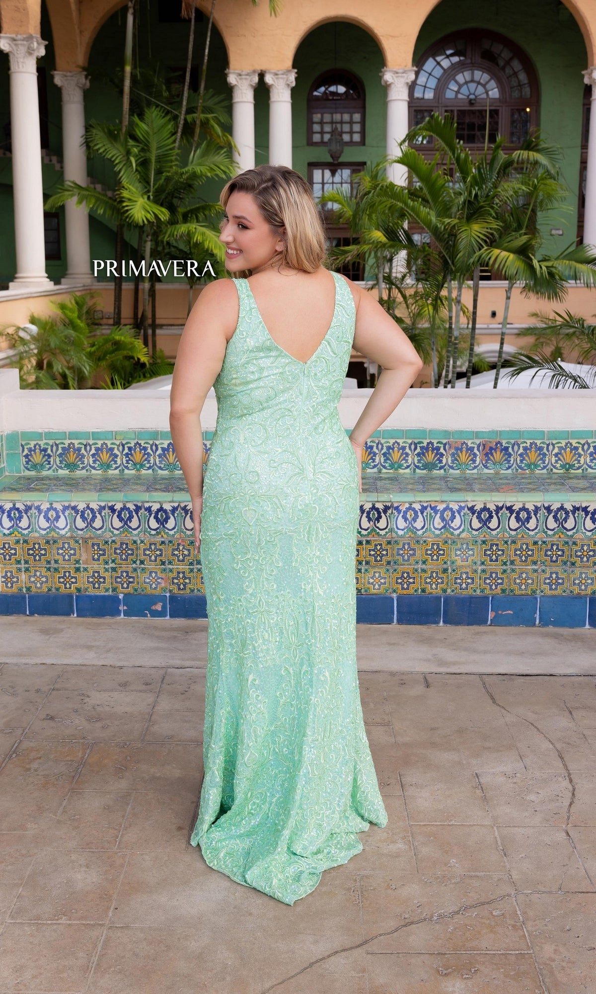 A woman in the Primavera Plus-Size V-Neck Beaded Prom Dress 14048, in light green, stands with her back to the camera in a tiled outdoor courtyard filled with palm plants and arches.